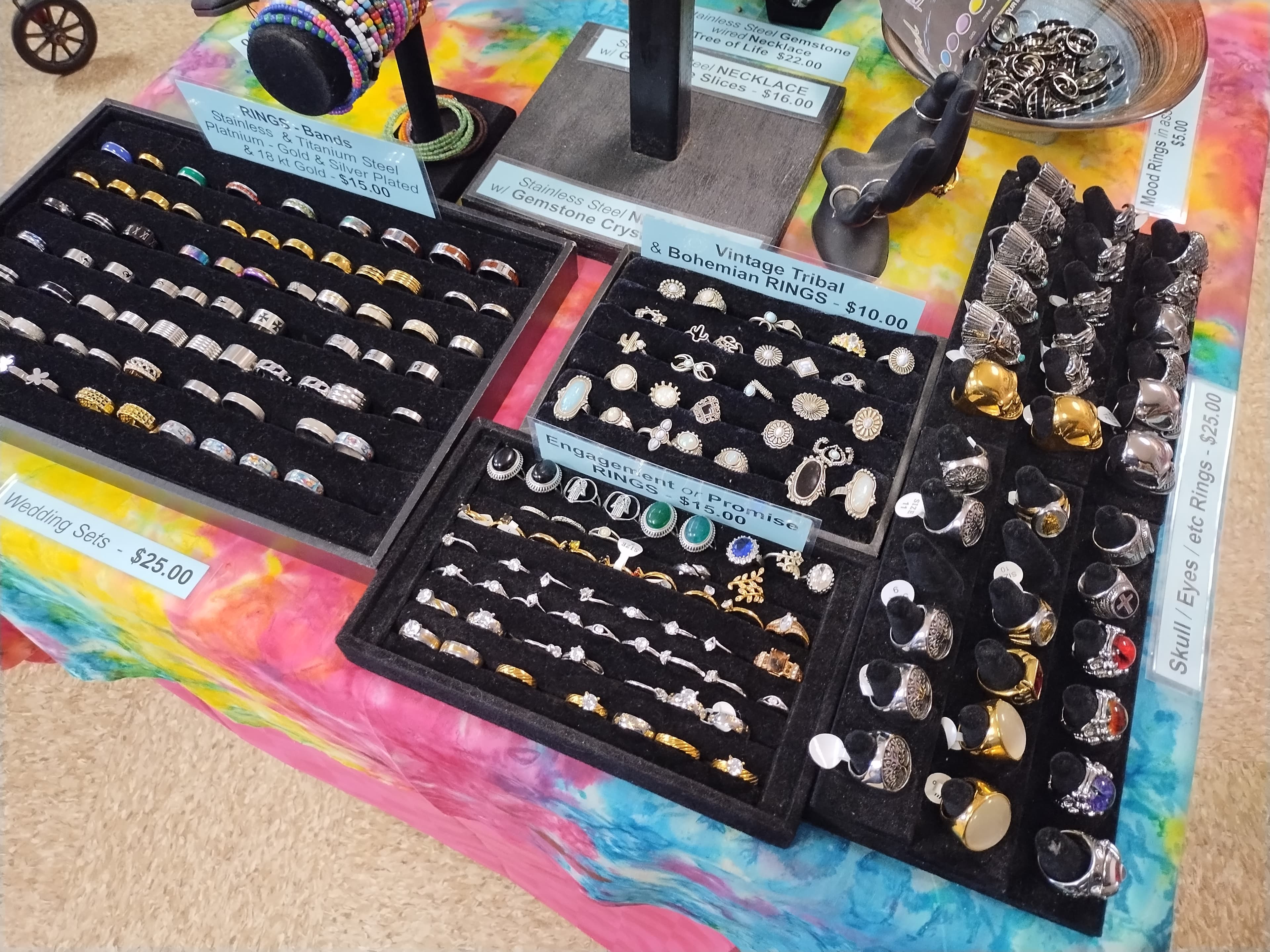 Assorted rings in black trays displayed on a vibrant tie-dye tablecloth at a jewelry stand.