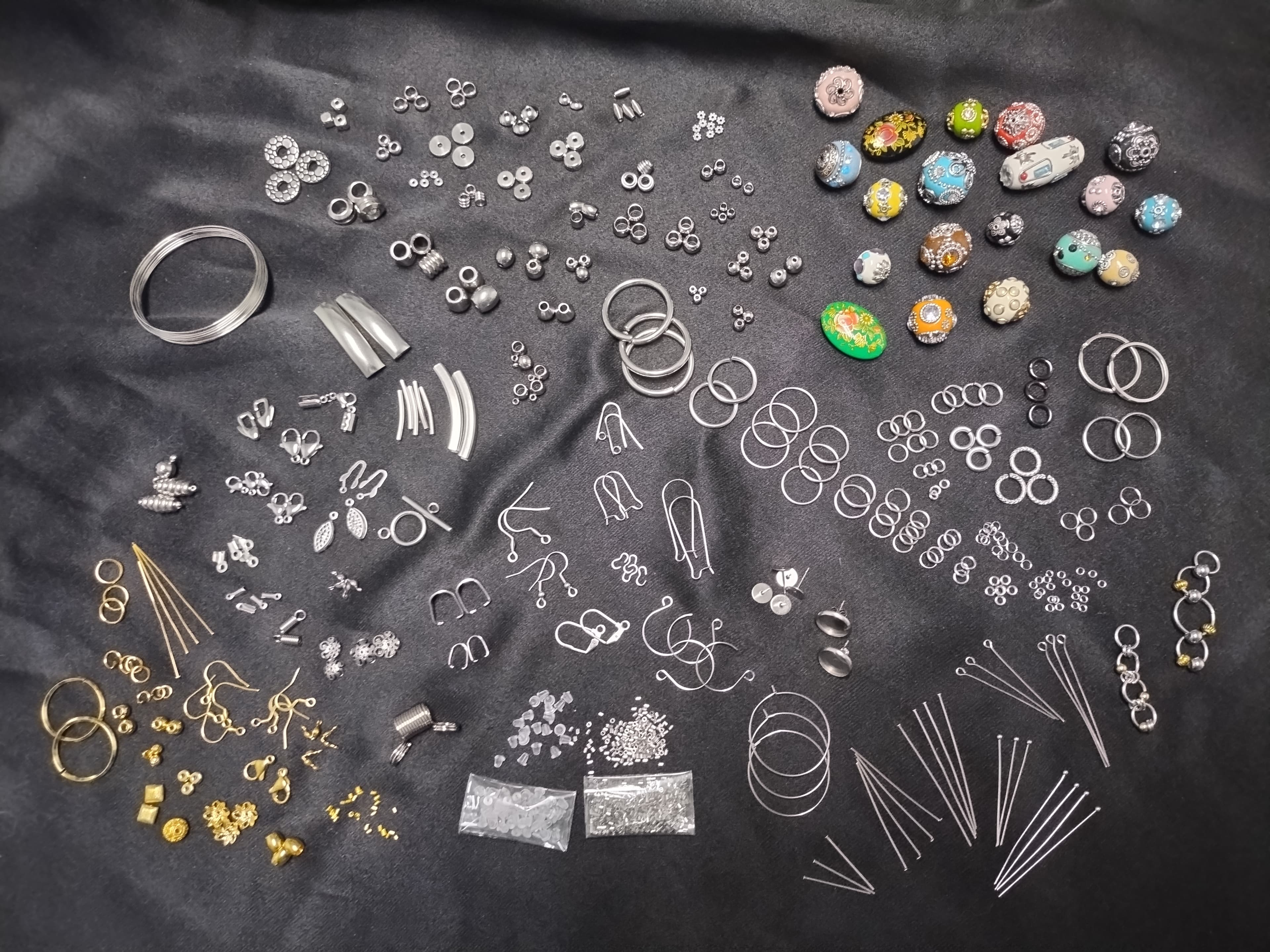 Various metal jewelry-making supplies, findings, and colorful beads scattered across a dark fabric surface.