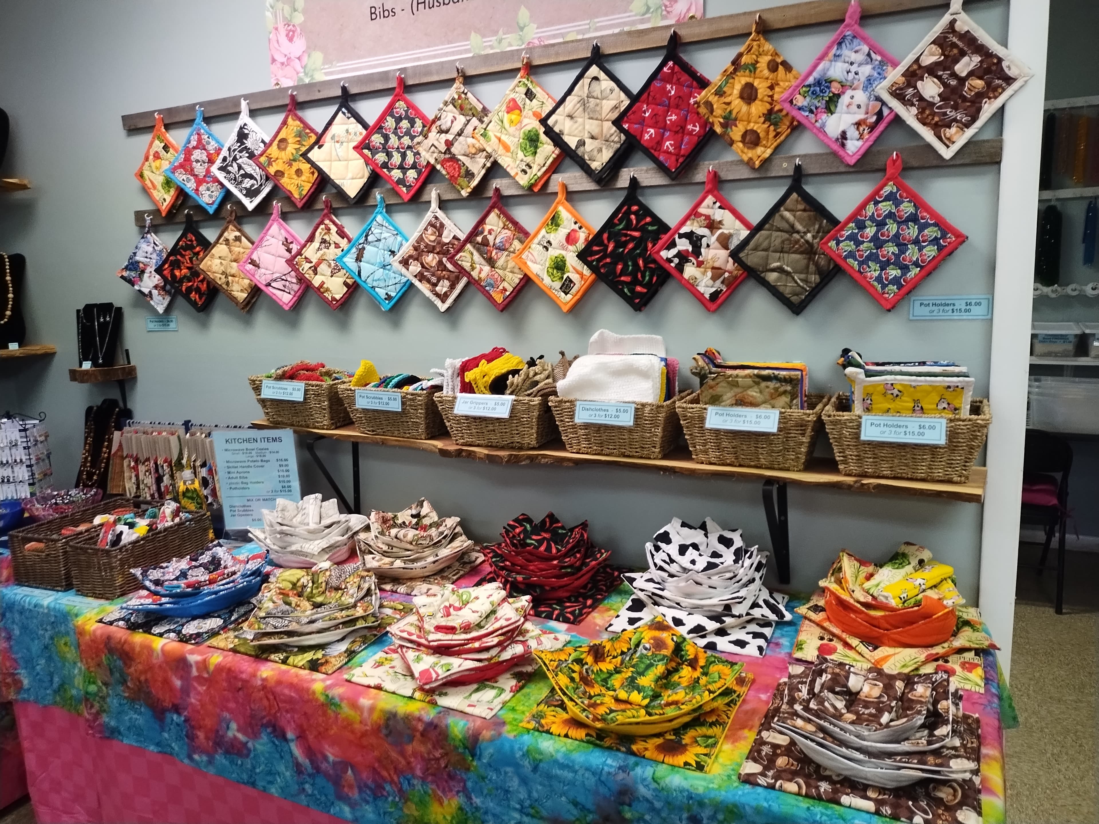 Colorful handmade pot holders and fabric bowl cozies displayed on shelves and a tie-dye table.