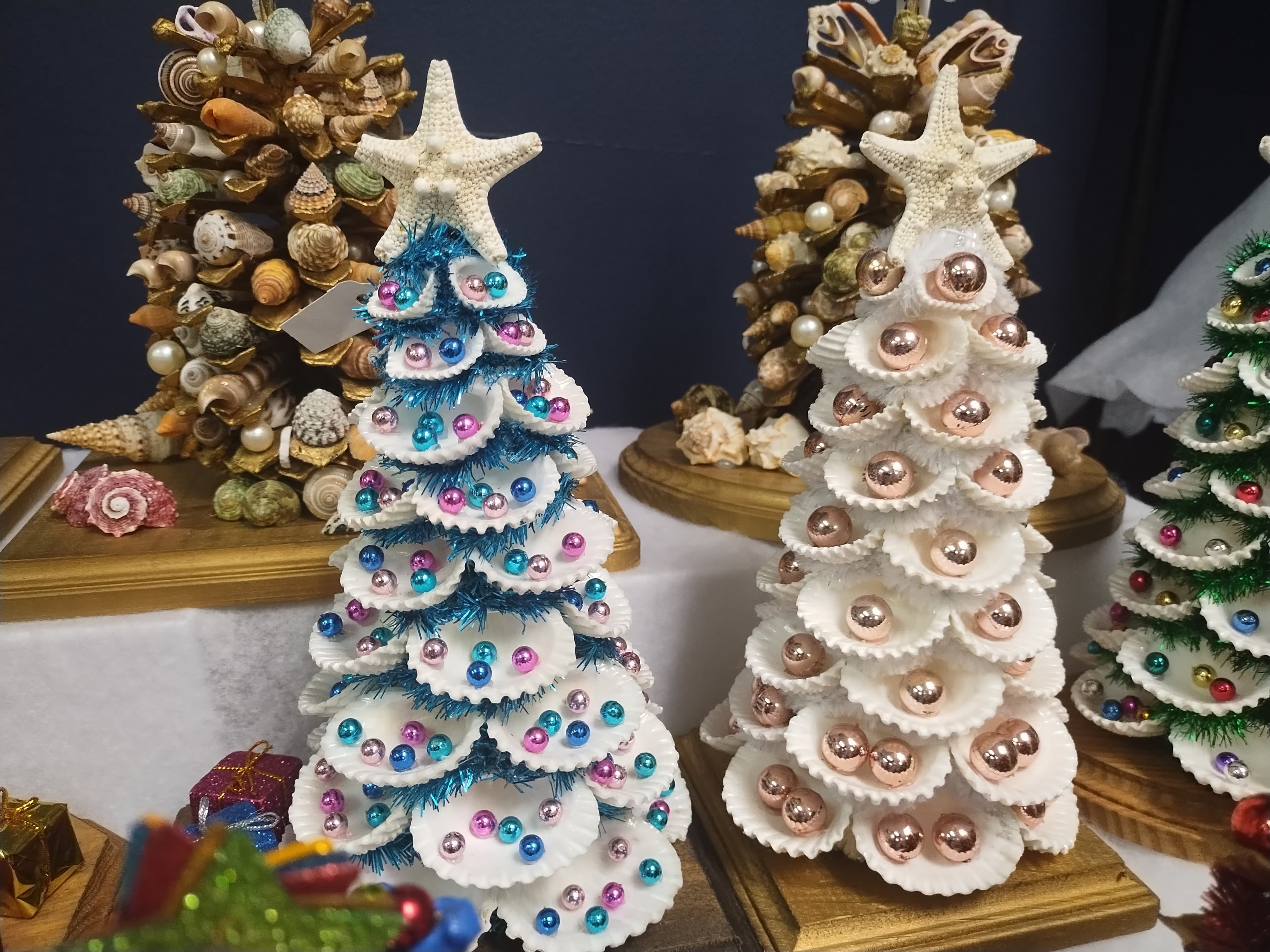 Miniature Christmas trees crafted from white seashells, featuring starfish toppers and colorful metallic ball ornaments.