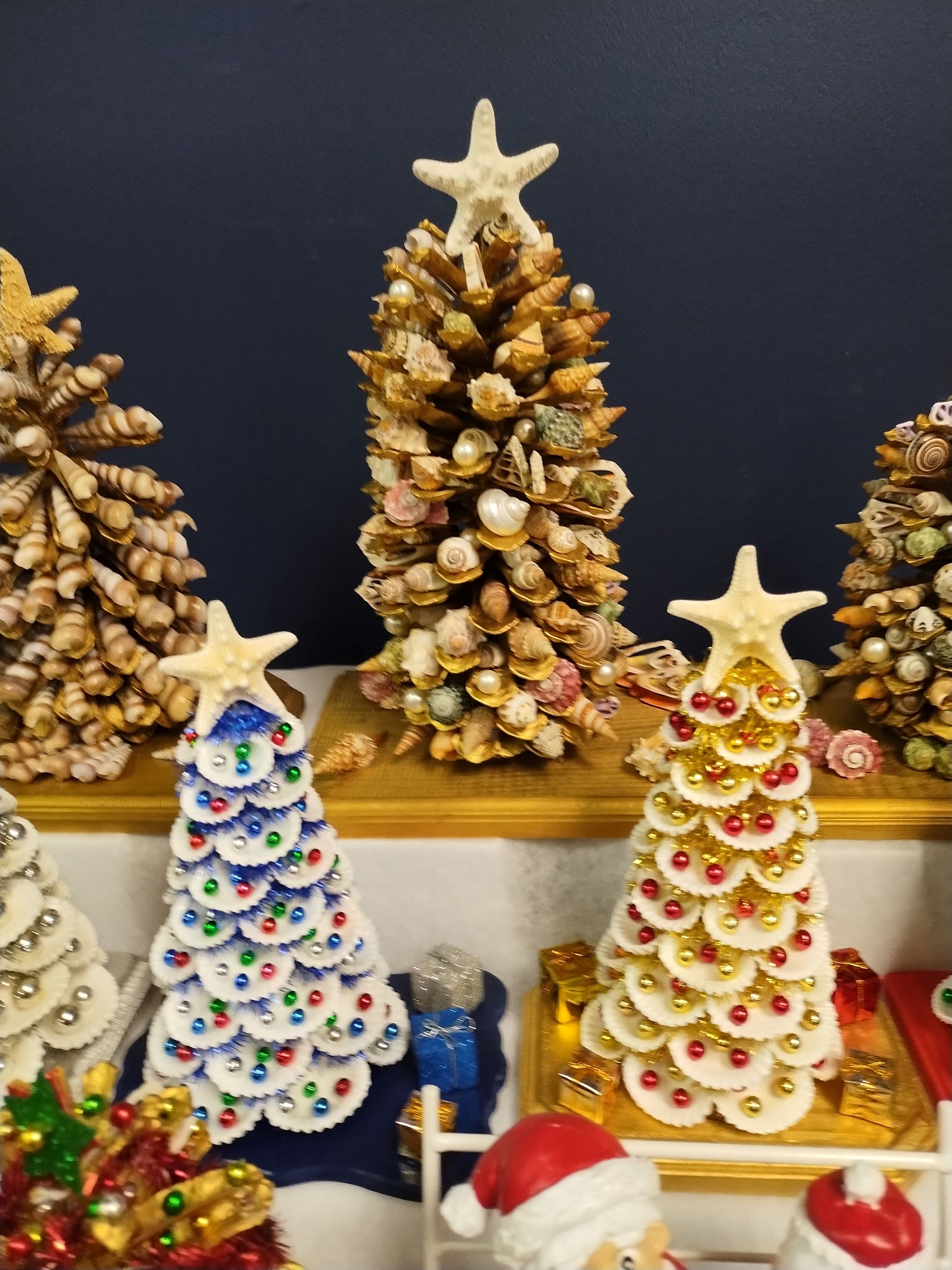 Miniature Christmas trees crafted from seashells and starfish, decorated with colorful beads on a shelf.